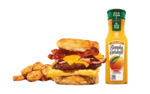 A fully loaded breakfast biscuit sandwich with multiple meats, cheese, and egg, served with juice and hashbrowns.
